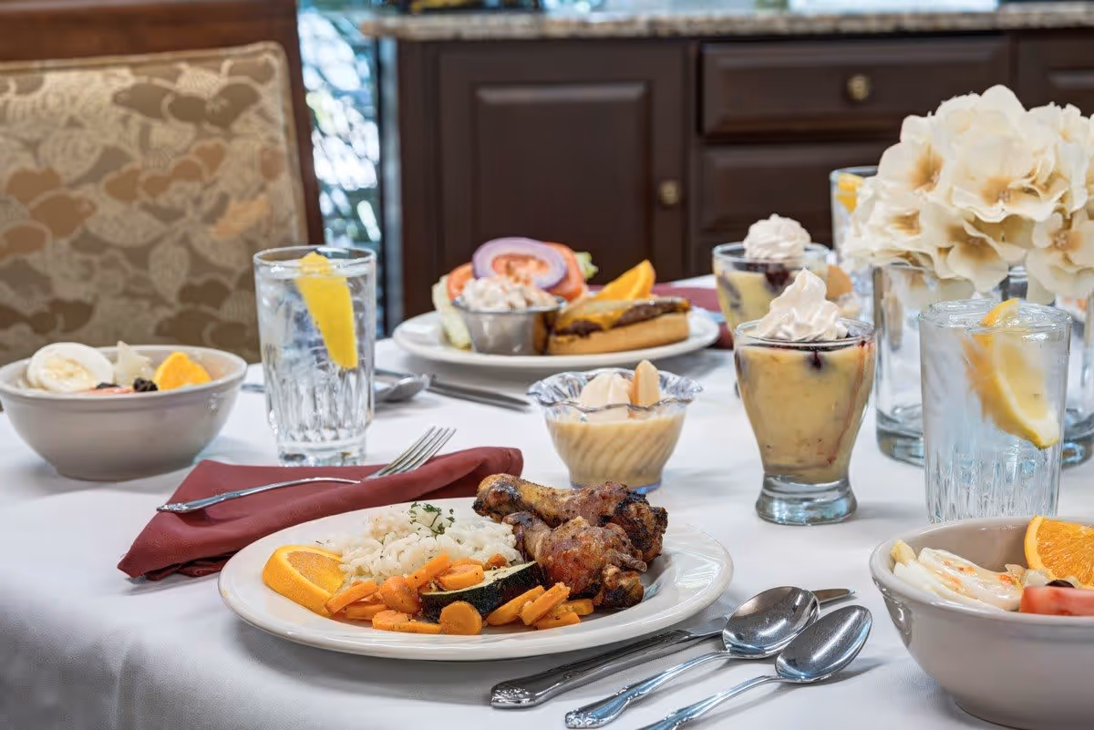 A dining table set with plates of food including grilled chicken drumsticks with rice and vegetables, a sandwich with lettuce and tomato, bowls of salad, glasses of water with lemon slices, and dessert cups topped with whipped cream. The table is covered with a white tablecloth and has a floral centerpiece. A cushioned chair with a patterned fabric is visible in the background.