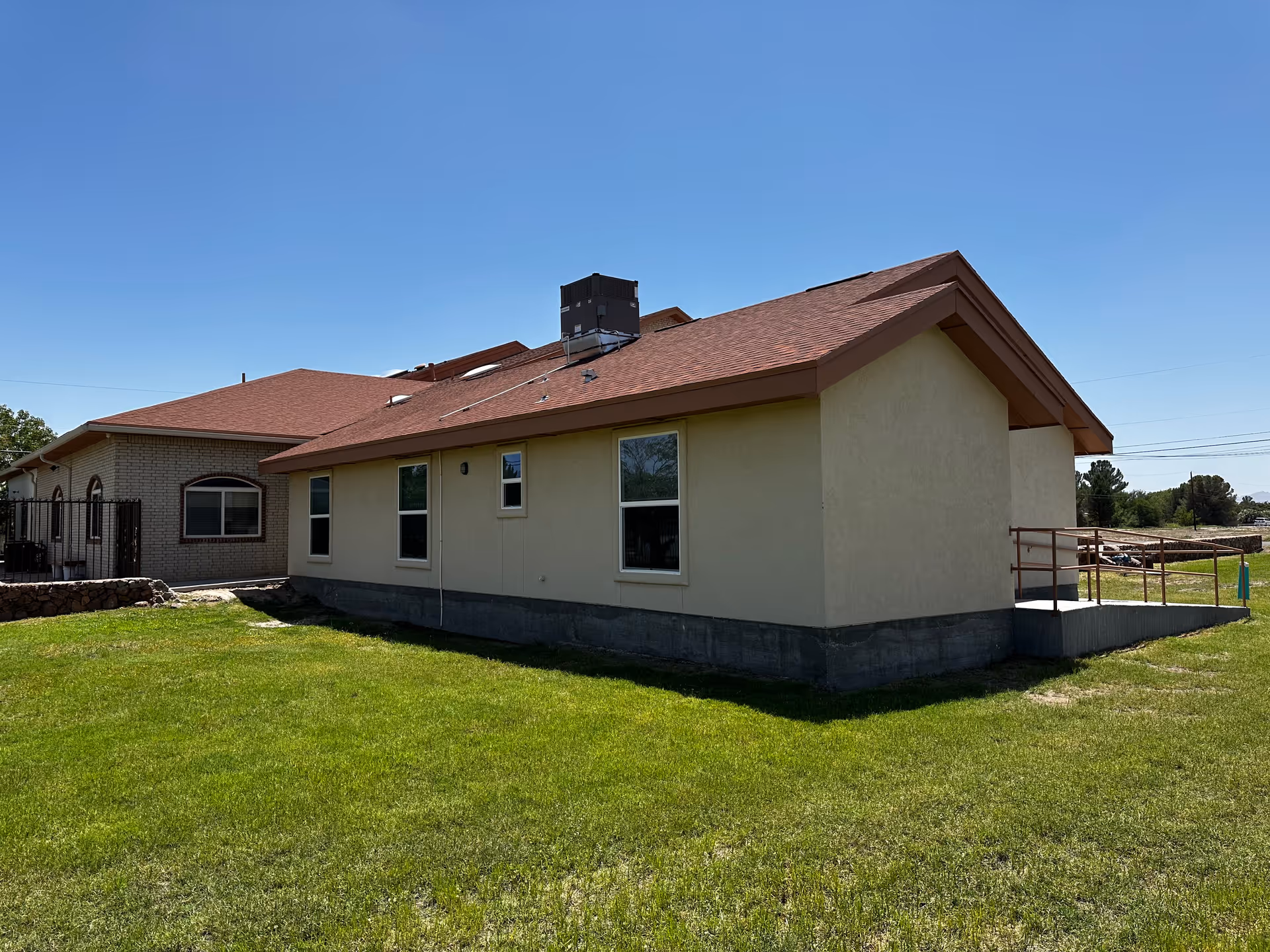 Single-story light-colored senior care building with a red roof, windows, an accessible ramp and a grassy yard under a clear blue sky.