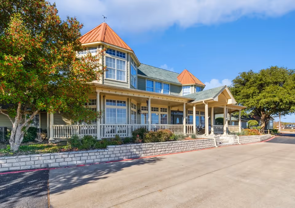 Exterior view of a senior living facility building with a porch, large windows, and two peaked roof sections with red-orange tops. There are trees and shrubs around the building, and a paved driveway in front under a partly cloudy blue sky.