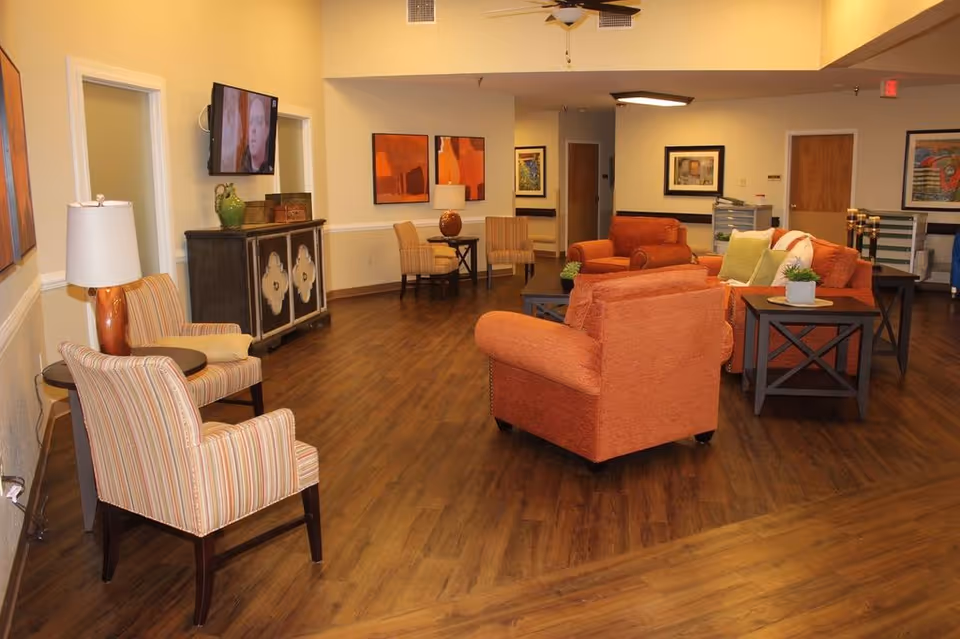 A spacious living room area in a nursing and rehabilitation center featuring wooden flooring, orange upholstered armchairs and sofas, striped accent chairs, side tables with lamps, wall-mounted TV, and framed artwork on the walls.
