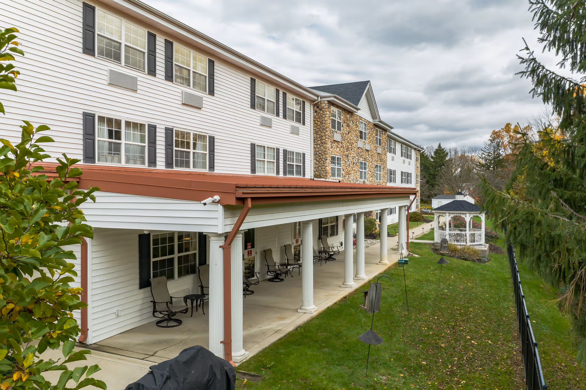 Exterior view of a three-story senior living building with a covered porch supported by columns and a gazebo on the lawn.