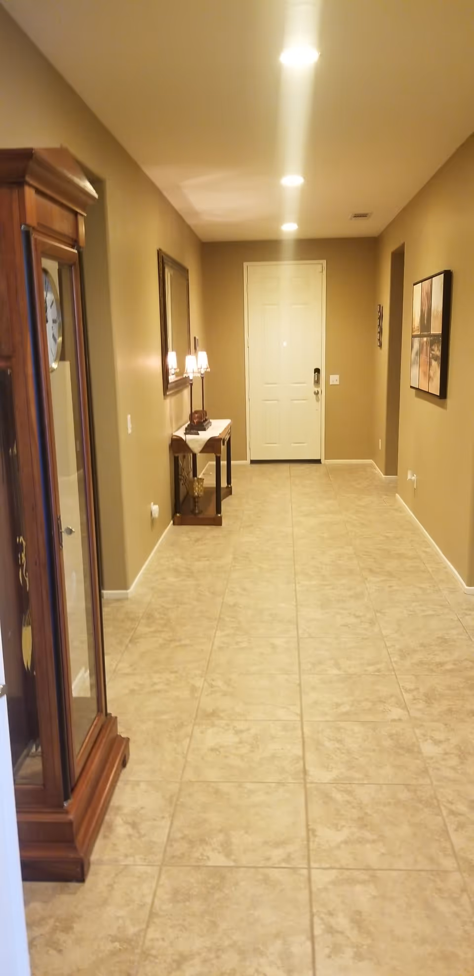 Long tiled entry hallway with a front door, side table and lamps, wall art, and a grandfather clock.