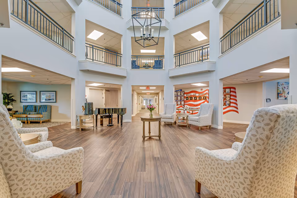 A spacious and well-lit senior living facility common area with high ceilings and a large hanging light fixture. The room features wood flooring, several upholstered armchairs arranged around a small round table with a flower vase, a black grand piano, and a hallway leading further into the building. The walls are decorated with patriotic-themed artwork and framed pictures.