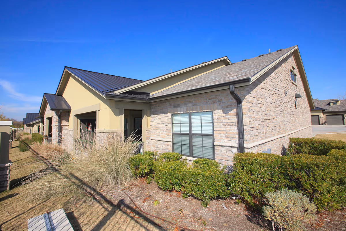 Exterior of a single-story stone-and-stucco residential building with windows, landscaping, and a clear blue sky.