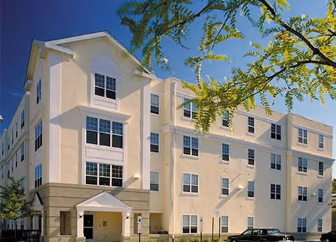 Exterior view of a multi-story beige building with multiple windows and a small entrance canopy, set against a clear blue sky with some tree branches in the foreground.