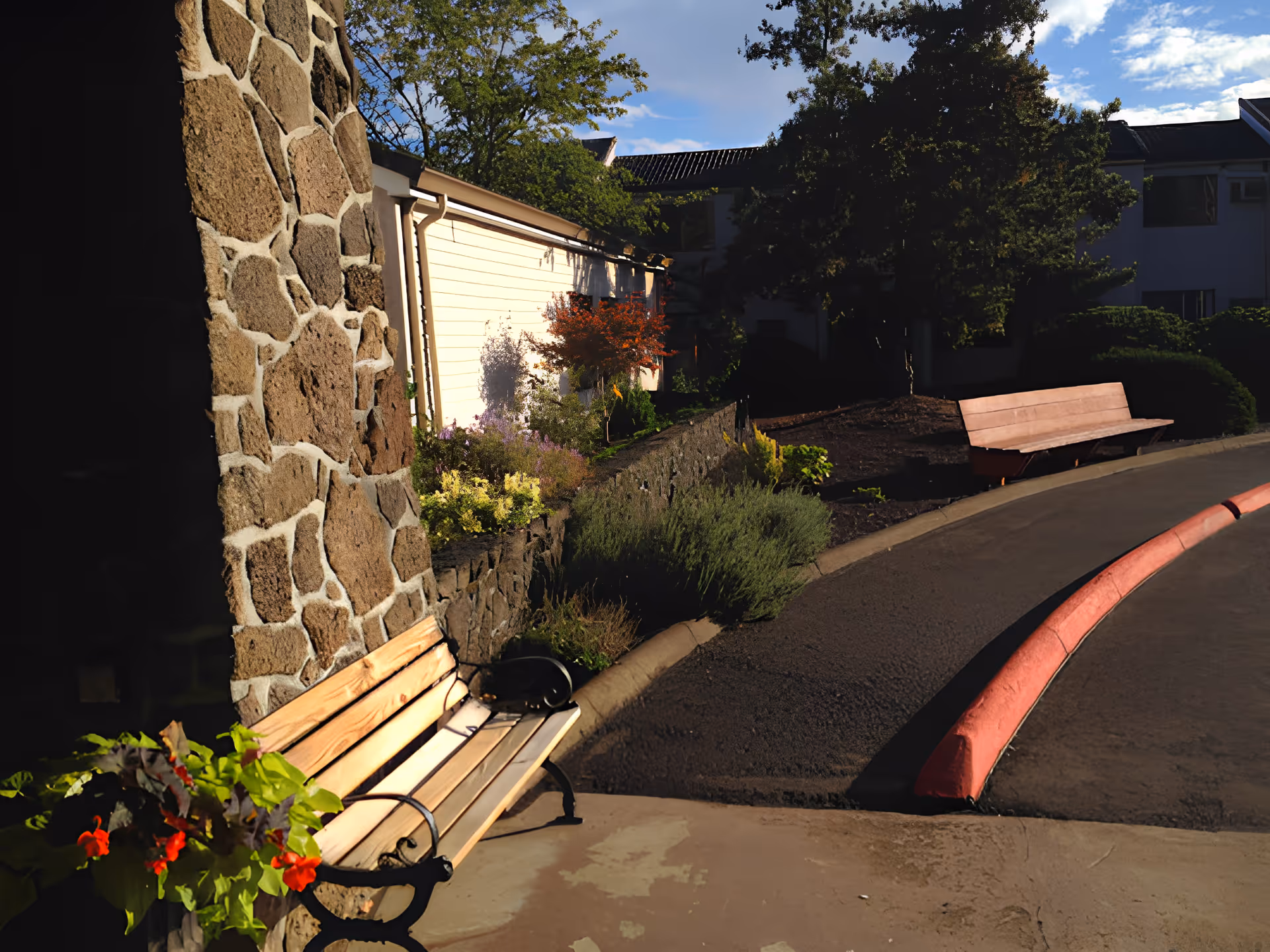 Outdoor area of Churchill Estates Retirement Community featuring two wooden benches along a curved paved pathway, a stone wall with plants and flowers, and trees providing shade under a partly cloudy sky.