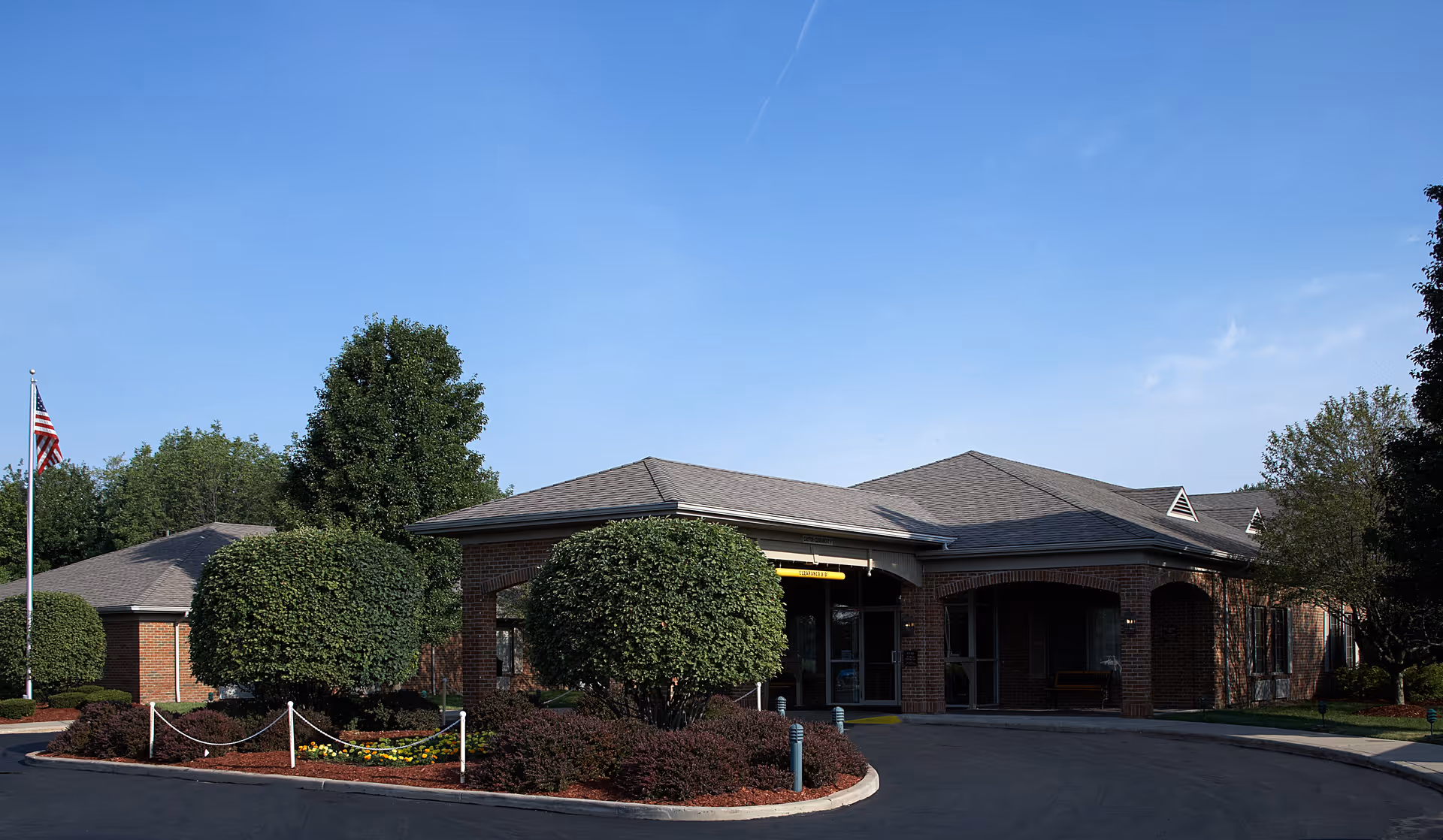 A single-story brick senior living facility with a curved driveway, manicured shrubs, and an American flag near the entrance.