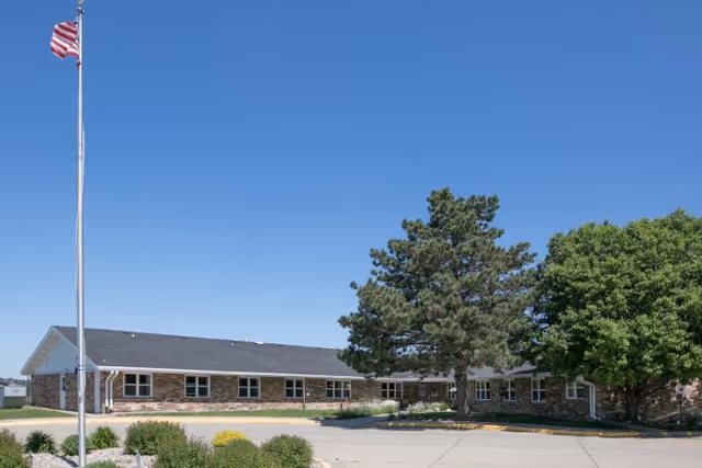 Single-story brick building with multiple windows, surrounded by trees and shrubs under a clear blue sky. An American flag on a tall flagpole is visible on the left side of the image. The building appears to be part of a senior living facility.