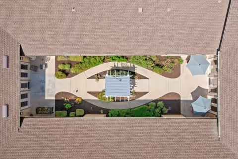 Aerial view of a rectangular courtyard garden enclosed by a building roof. The courtyard features a central pergola with benches, curved walkways, landscaped greenery, and two patio umbrellas with seating areas on one end.