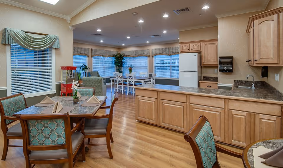 Interior view of a senior living facility dining and common area with wooden flooring, multiple tables with chairs, a kitchen counter with cabinets, a white refrigerator, a popcorn machine, and large windows with blinds and valances letting in natural light.