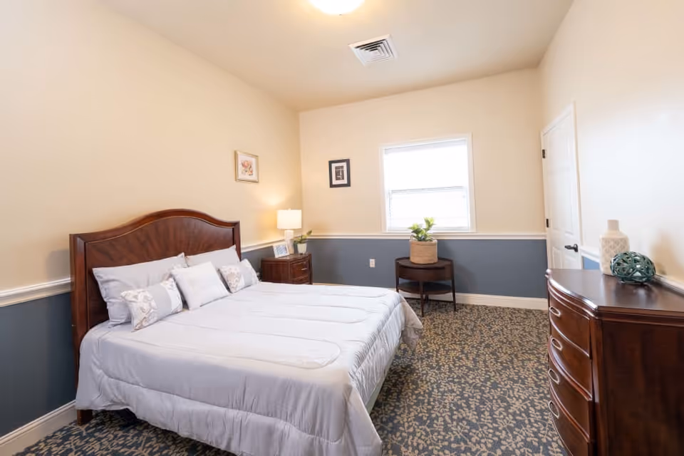 A neatly made bed with white bedding and multiple pillows in a bedroom with beige and blue walls. There is a wooden nightstand with a lamp and picture frame beside the bed, a small round table with a plant near the window, and a wooden dresser with decorative items on top. The room has patterned carpet flooring and a closed white door.