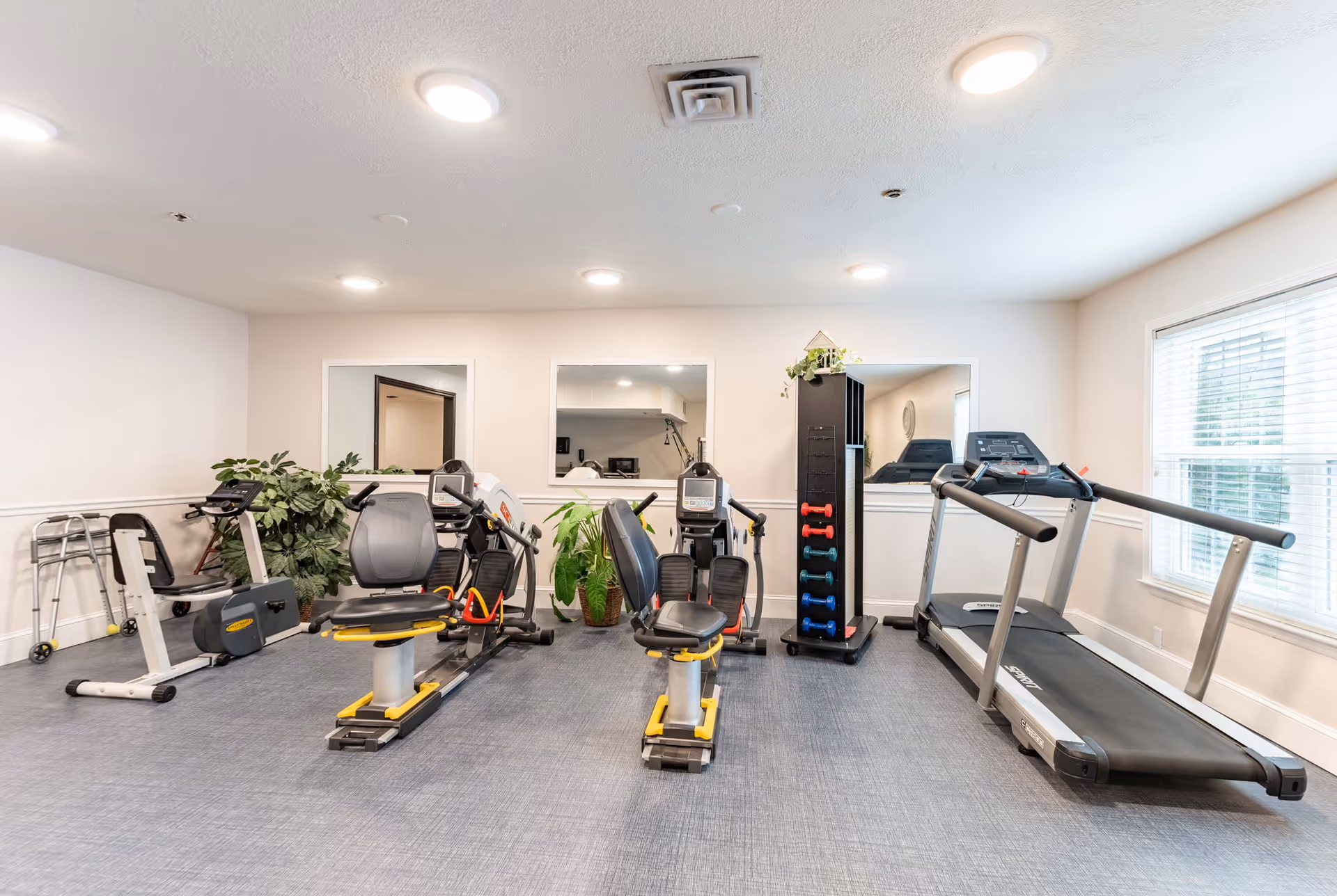 A senior living facility exercise room with various fitness equipment including two recumbent exercise bikes, a treadmill, a rack of colorful dumbbells, and a walker. The room has light-colored walls, three large windows, and ceiling lights providing bright illumination.