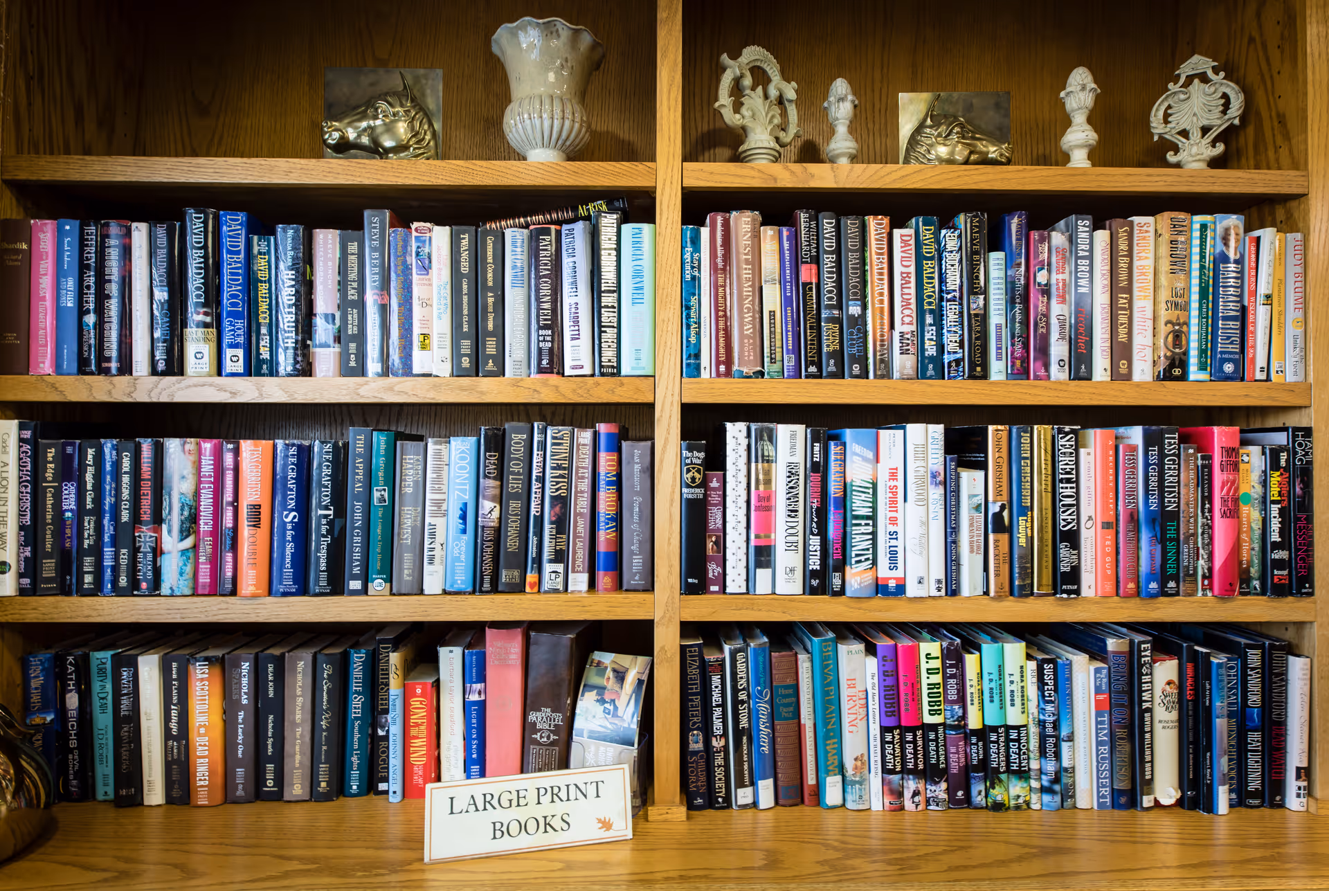 Wooden bookshelf filled with multiple rows of large print books, with decorative ceramic figurines and vases on the top shelf. A small sign on the bottom shelf reads 'LARGE PRINT BOOKS'.
