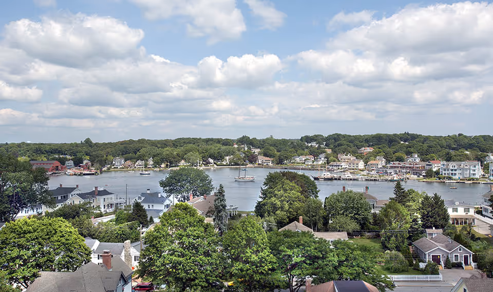 A scenic view of a waterfront town with houses, trees, and boats on the water under a partly cloudy sky.