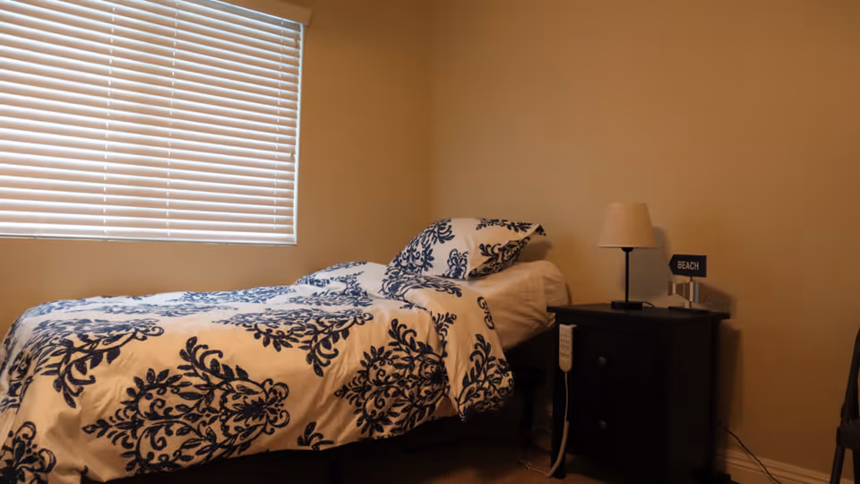 A small bedroom with a single bed covered in white bedding with dark blue floral patterns. Next to the bed is a black nightstand with two drawers, a beige lampshade on a black lamp, and a small sign that says 'BEACH'. A window with closed white blinds is on the left wall, and the walls are painted beige.
