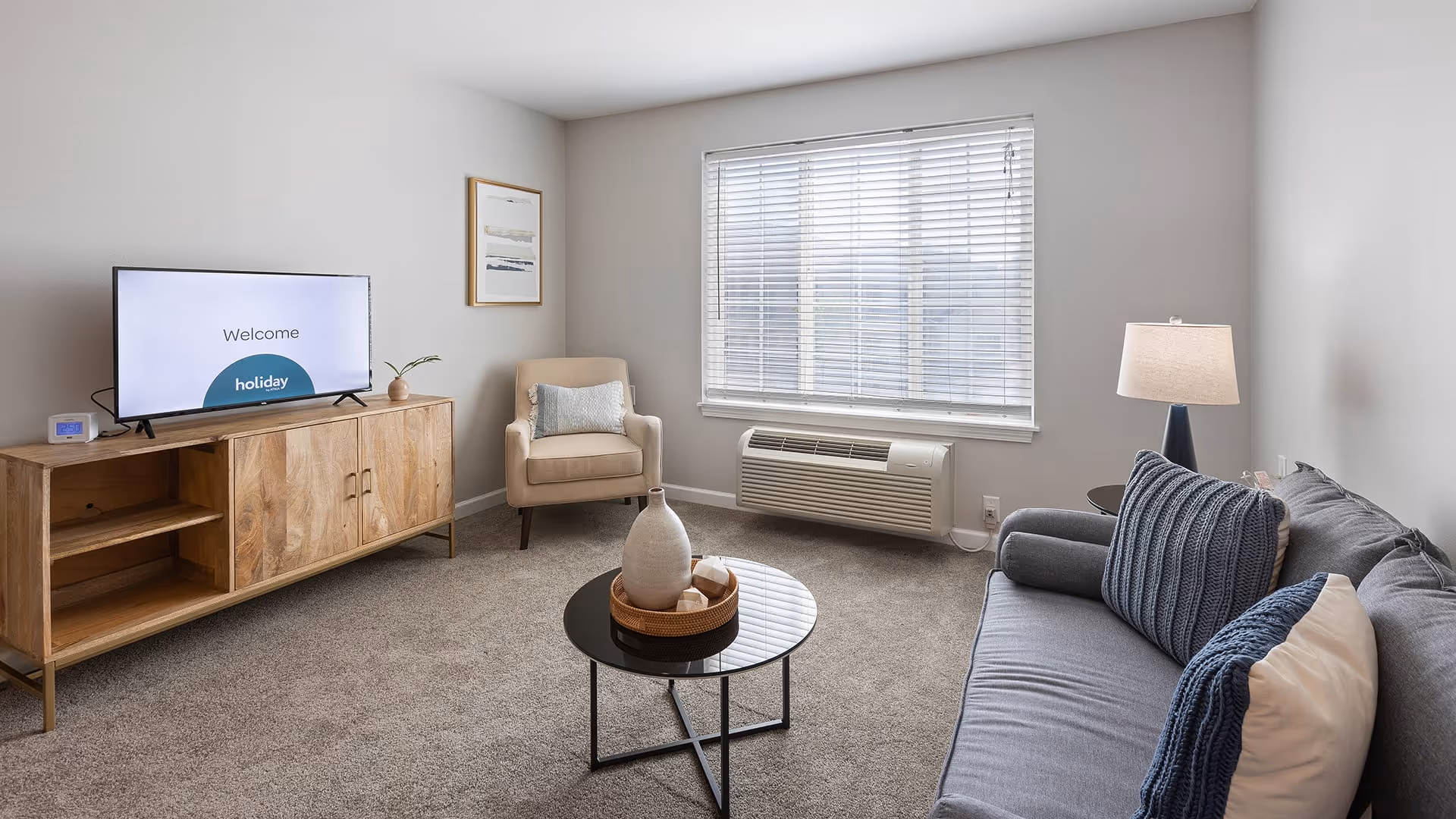 A cozy living room with a gray sofa adorned with blue and white pillows, a beige armchair with a decorative pillow, a wooden TV stand with a flat-screen TV displaying a welcome message, a round black coffee table with decorative items, a table lamp, and a large window with white blinds letting in natural light.