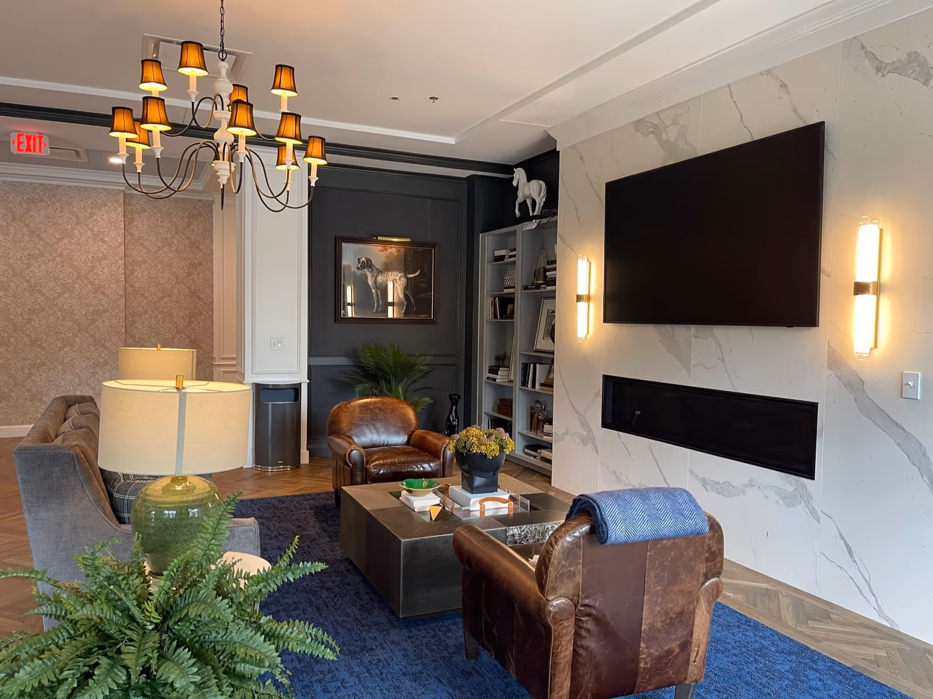 A cozy living room with two brown leather armchairs, a gray sofa, a large flat-screen TV mounted on a white marble wall above a modern fireplace, a dark blue rug, a coffee table with books and a flower pot, a green table lamp, a chandelier with multiple small lampshades, a bookshelf filled with books and decor, and a framed picture of a dog on a dark accent wall.