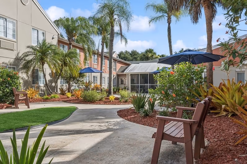 Outdoor courtyard area at The Gardens of Port St. Lucie featuring a paved walkway, brown wooden benches, tropical plants, palm trees, and blue patio umbrellas under a partly cloudy sky.