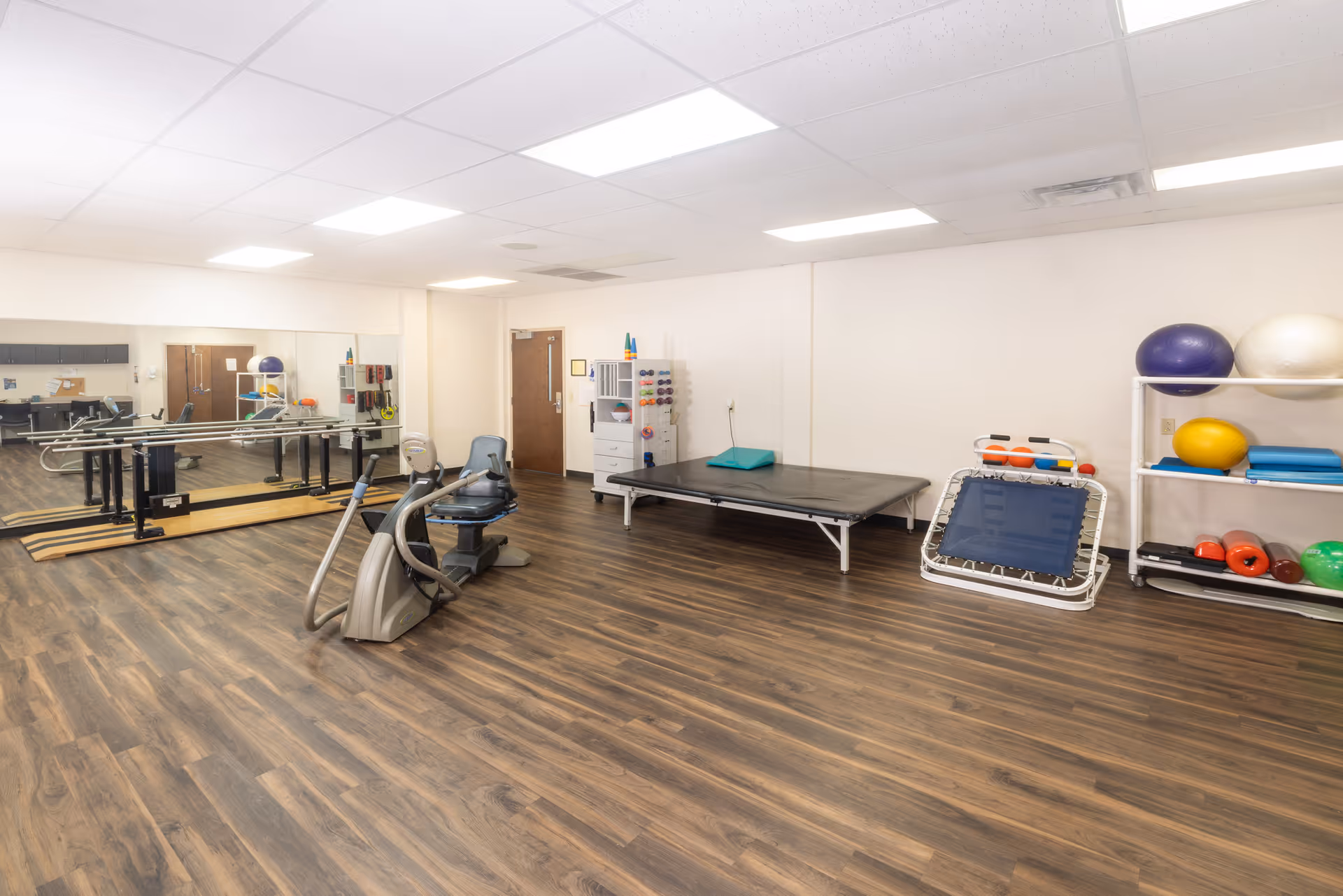 A spacious physical therapy or rehabilitation room with wooden flooring, exercise equipment including a stationary bike, parallel bars, a therapy table, a small trampoline, and shelves holding various exercise balls and weights. The room has white walls, a ceiling with fluorescent lights, and a large mirror along one wall.