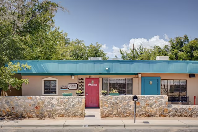 Exterior front view of a single-story building with a stone wall and a red door that has a 'Welcome' sign. The building has a teal roof trim and several windows with blinds. There are trees and a clear blue sky in the background.