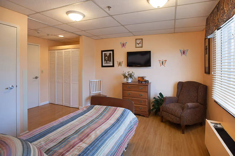 A cozy bedroom in a nursing and rehab center featuring a bed with a striped blanket, a wooden dresser with decorative items, a patterned armchair, a white chair, and a wall-mounted TV. The walls are decorated with butterfly decals and framed pictures. There is a window with blinds and a valance, and the floor is wooden.