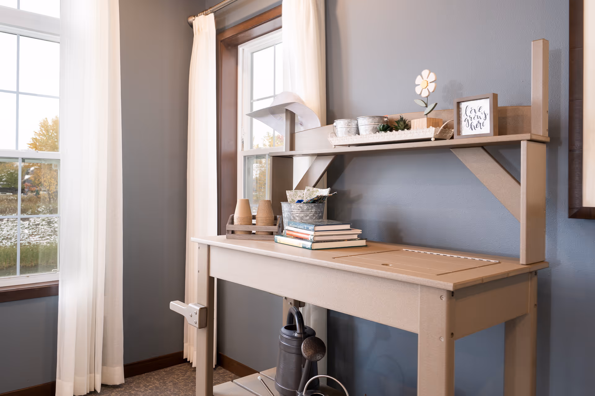A beige wooden potting bench with gardening supplies including small pots, a watering can, books, and decorative items, positioned against a gray wall with two windows framed by white curtains allowing natural light into the room.