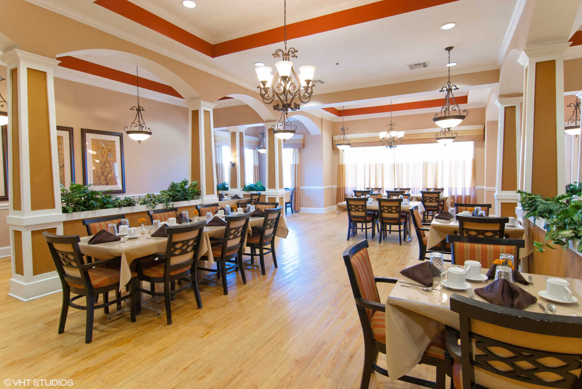 Spacious dining room with multiple set tables and chairs, chandeliers, and warm-colored decor.