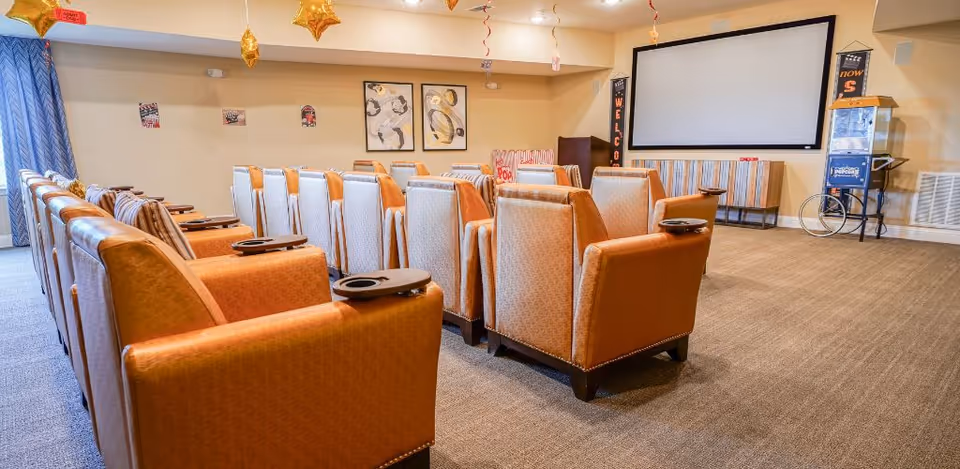 Rows of upholstered theater-style chairs facing a large projection screen in a cozy media room with balloons and a popcorn machine.