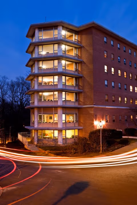 Exterior view of a multi-story brick building at dusk with large windows showing illuminated interior rooms. Light trails from passing cars curve around the building, and a street lamp is visible near the entrance.