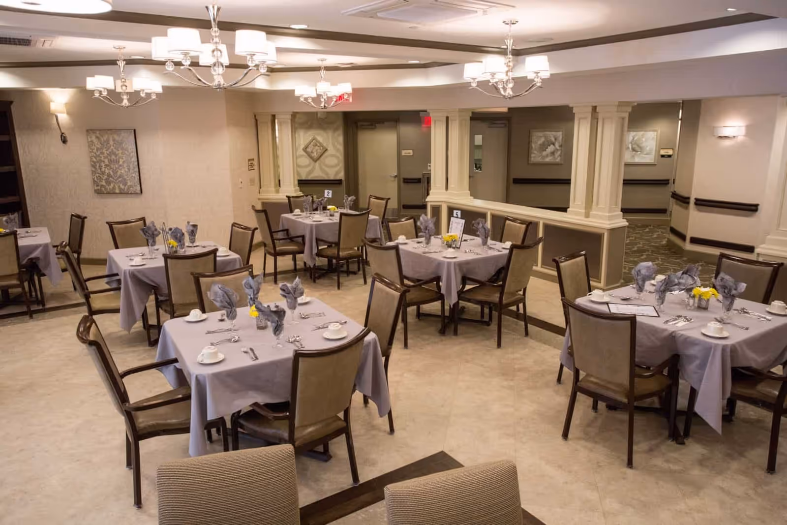 A dining room in an assisted living facility with several square tables covered with light purple tablecloths. Each table is set with cups, saucers, silverware, and folded napkins, and some tables have small yellow flower centerpieces. The room has beige walls, decorative wall art, and elegant chandeliers hanging from the ceiling. There are multiple chairs around each table and a large mirror on one wall reflecting the room.