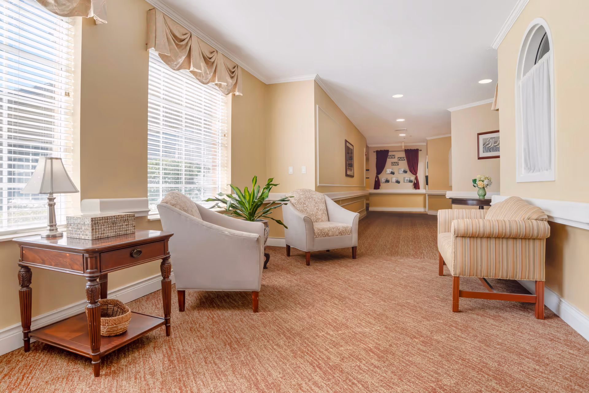 A bright and spacious hallway in a senior living facility with beige walls and carpeted floor. The hallway features large windows with blinds and valances on the left side, allowing natural light to fill the space. There are several upholstered chairs and a striped loveseat arranged along the walls, along with a wooden side table holding a lamp and a decorative box. A green potted plant and framed artwork add to the welcoming atmosphere.
