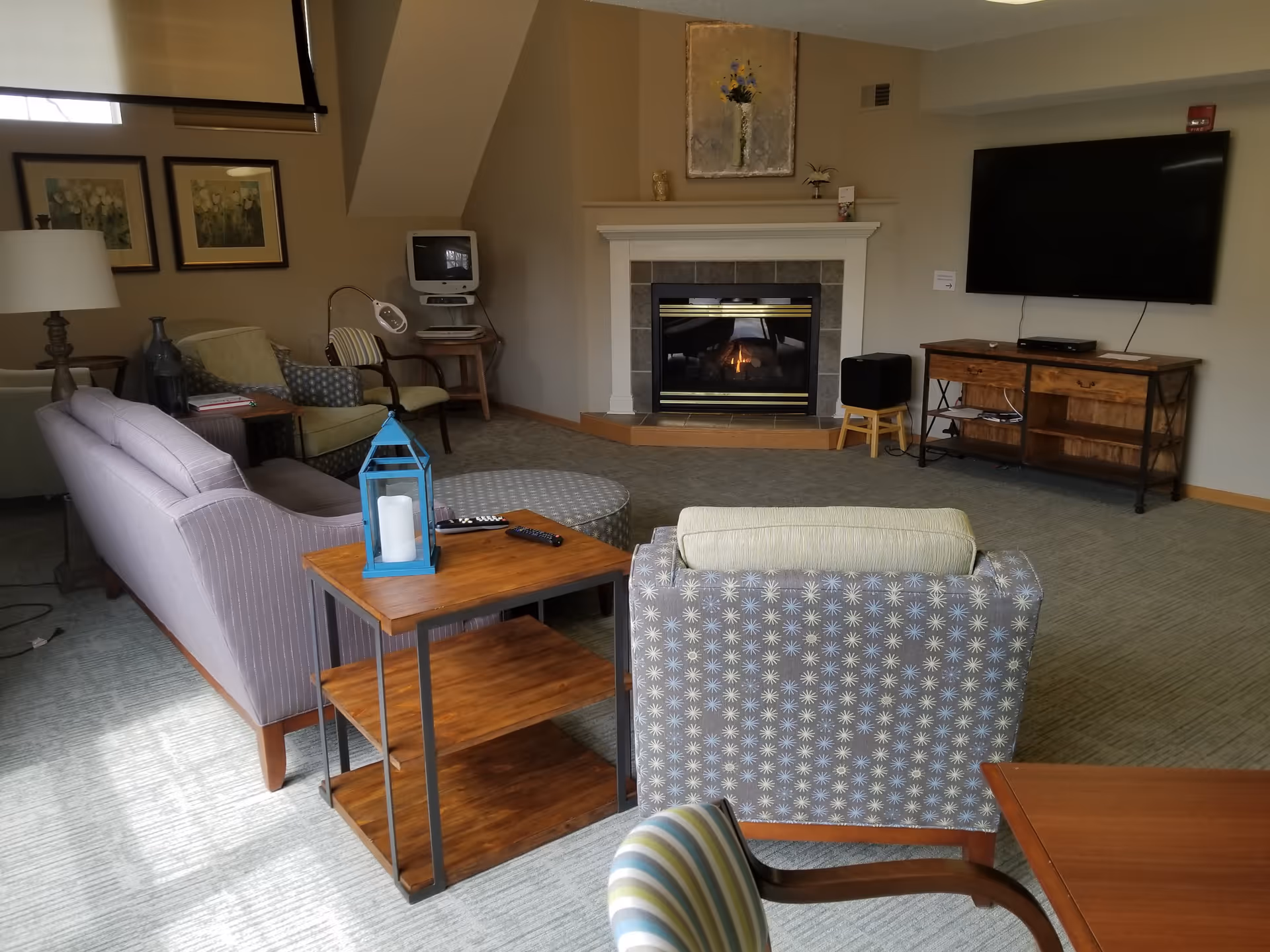 A cozy living room in a senior living facility featuring a fireplace with a lit fire, a large flat-screen TV mounted on the wall, a wooden TV stand, and various seating options including a patterned armchair, a striped sofa, and other chairs. The room is decorated with framed artwork, a table lamp, and a blue lantern candle holder on a wooden side table.