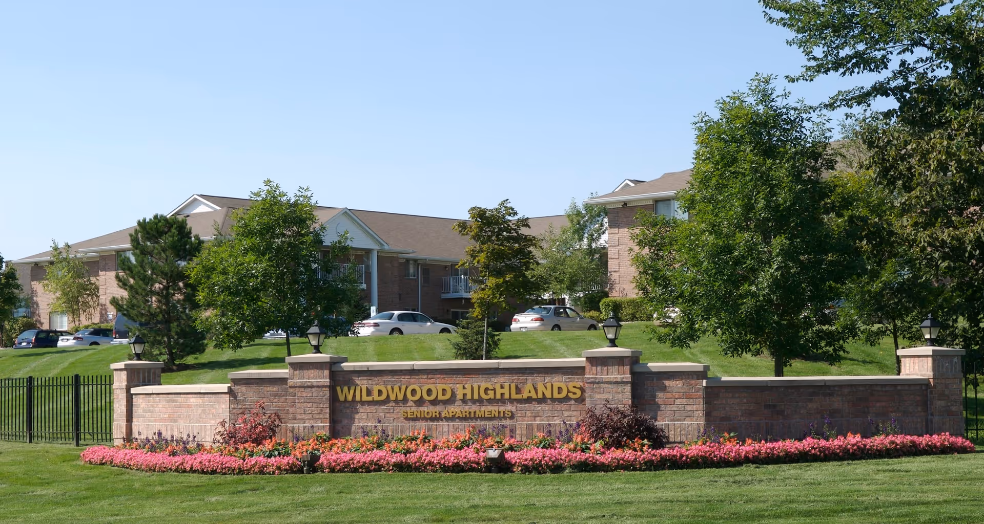Exterior view of Wildwood Highlands Senior Apartments with a brick sign in the foreground surrounded by pink flowers and green grass, and apartment buildings with parked cars and trees in the background under a clear blue sky.