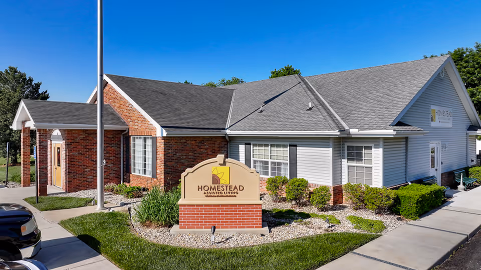Front exterior of Homestead Assisted Living building with a prominent entrance sign and landscaped grounds.