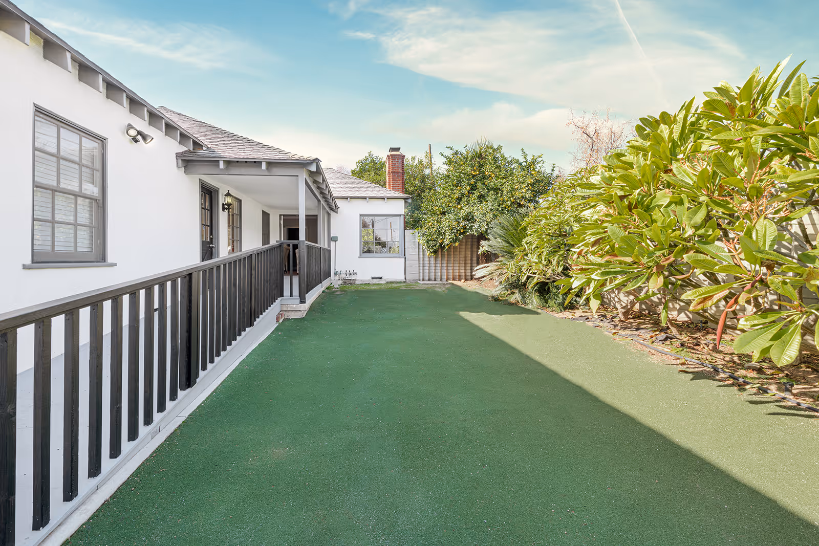 Outdoor view of a pathway alongside a white building with black railings and windows. The pathway is covered with green artificial turf and bordered by lush green bushes and trees under a partly cloudy sky.