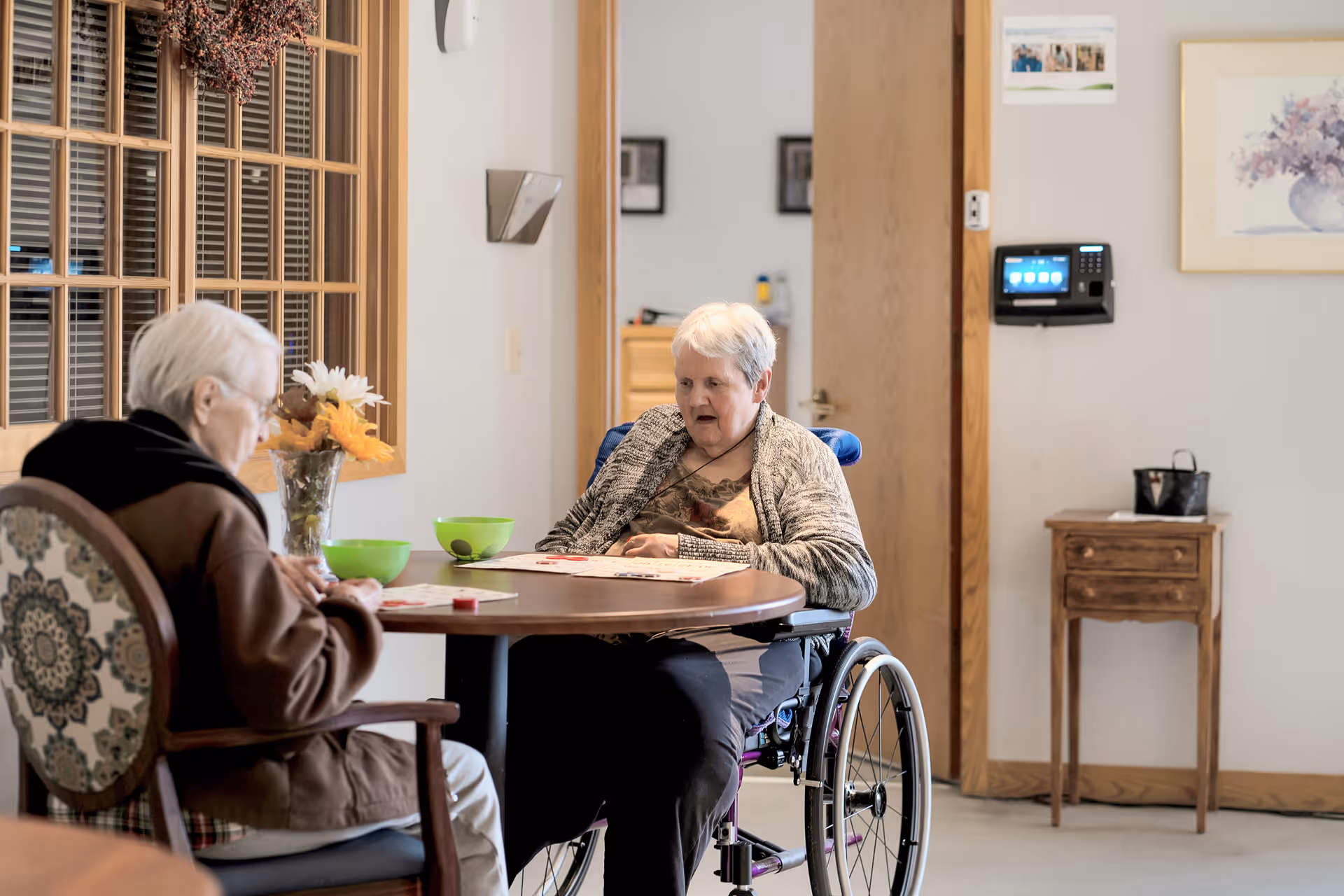 Two elderly women sitting at a round wooden table playing a game. One woman is in a wheelchair and the other is seated in a chair with a patterned backrest. There are green bowls and a vase with flowers on the table. The room has light-colored walls, a wooden door, and framed pictures on the wall.