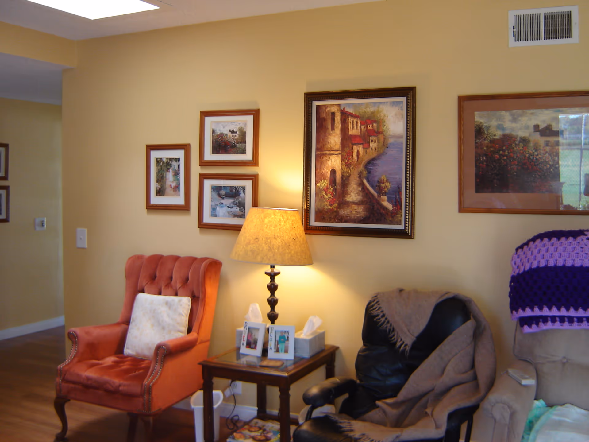 A cozy living room area with a red upholstered armchair, a black leather recliner draped with a brown blanket, and a beige armchair with a purple and pink crocheted blanket. A wooden side table with a lamp, tissue boxes, and framed photos sits between the chairs. The walls are painted yellow and decorated with several framed paintings and pictures.