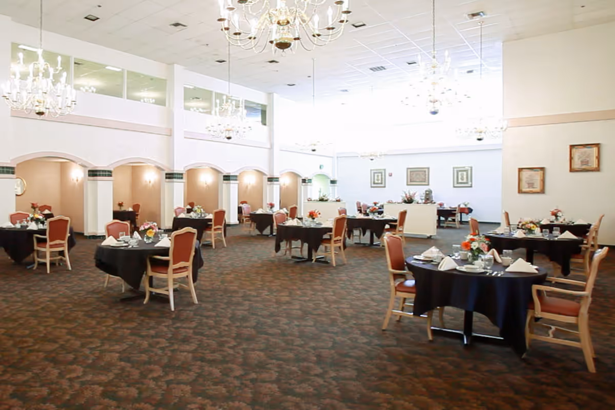 A spacious dining room with multiple round tables covered with black tablecloths, each set with white napkins, plates, and floral centerpieces. The room features high ceilings with several chandeliers and a patterned carpet. The walls have arches with lighting and framed artwork.