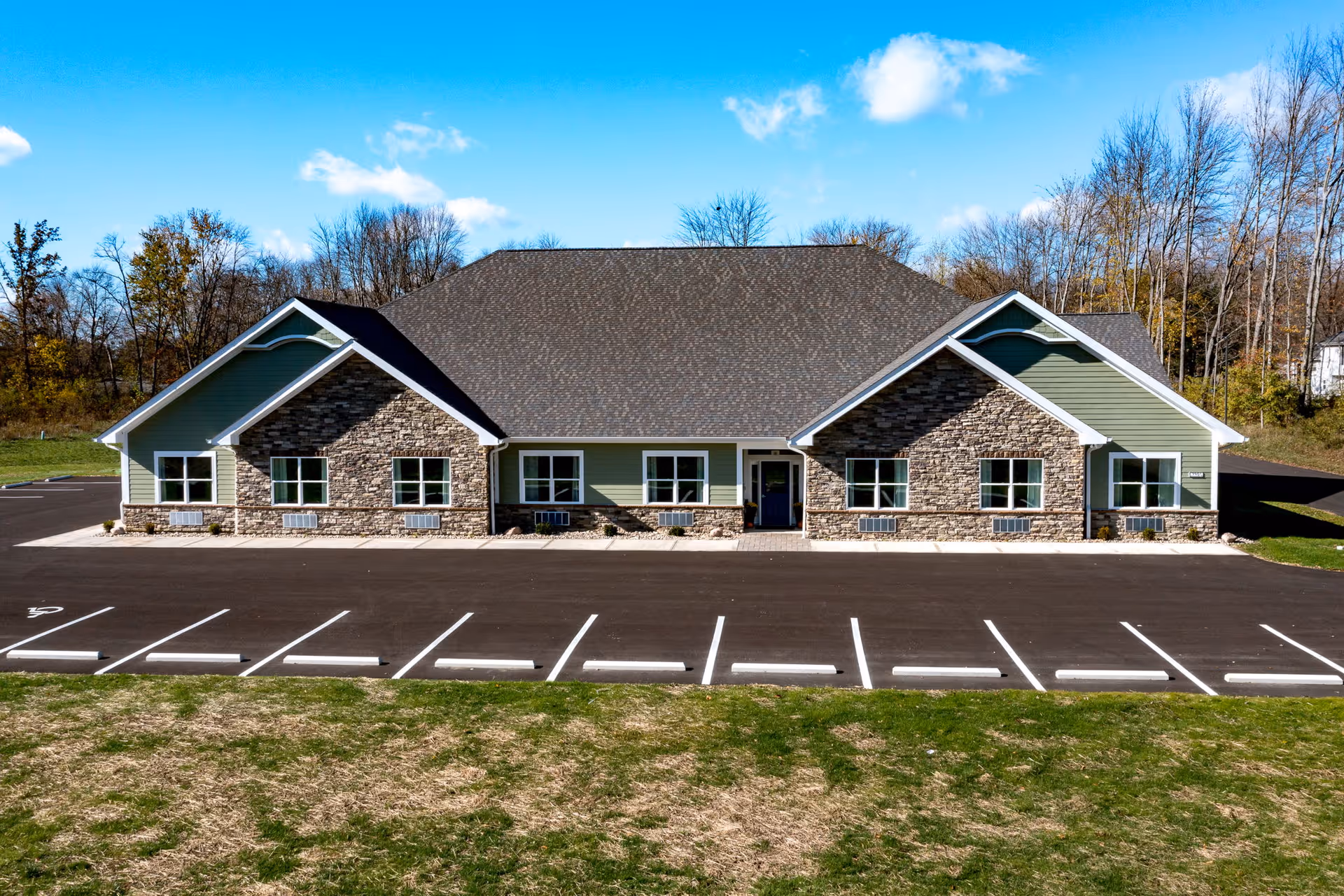 Single-story assisted living and memory care building with stone and green siding, rows of windows and an empty parking lot in front under a blue sky.