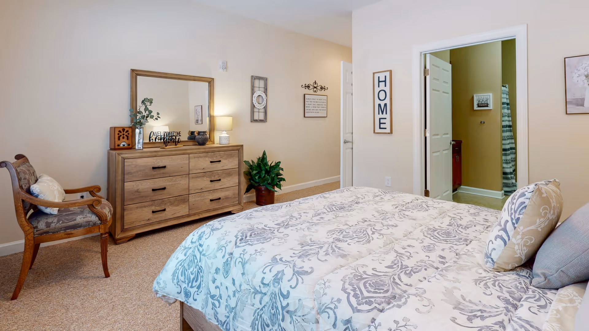 Bright furnished bedroom with a patterned bed, wooden dresser and chair, and an open door to an adjoining bathroom.