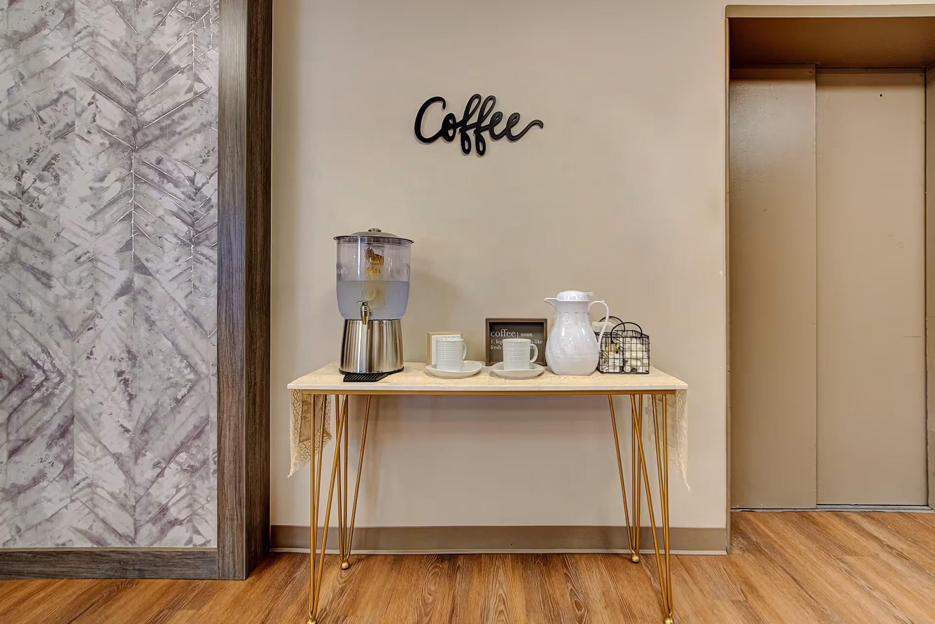A small coffee station with a water dispenser, two cups and saucers, a white pitcher, and a small wire basket on a table with gold legs. The wall behind has a decorative sign that says 'Coffee'. To the right is a closed elevator door, and to the left is a wall with a textured, patterned design.