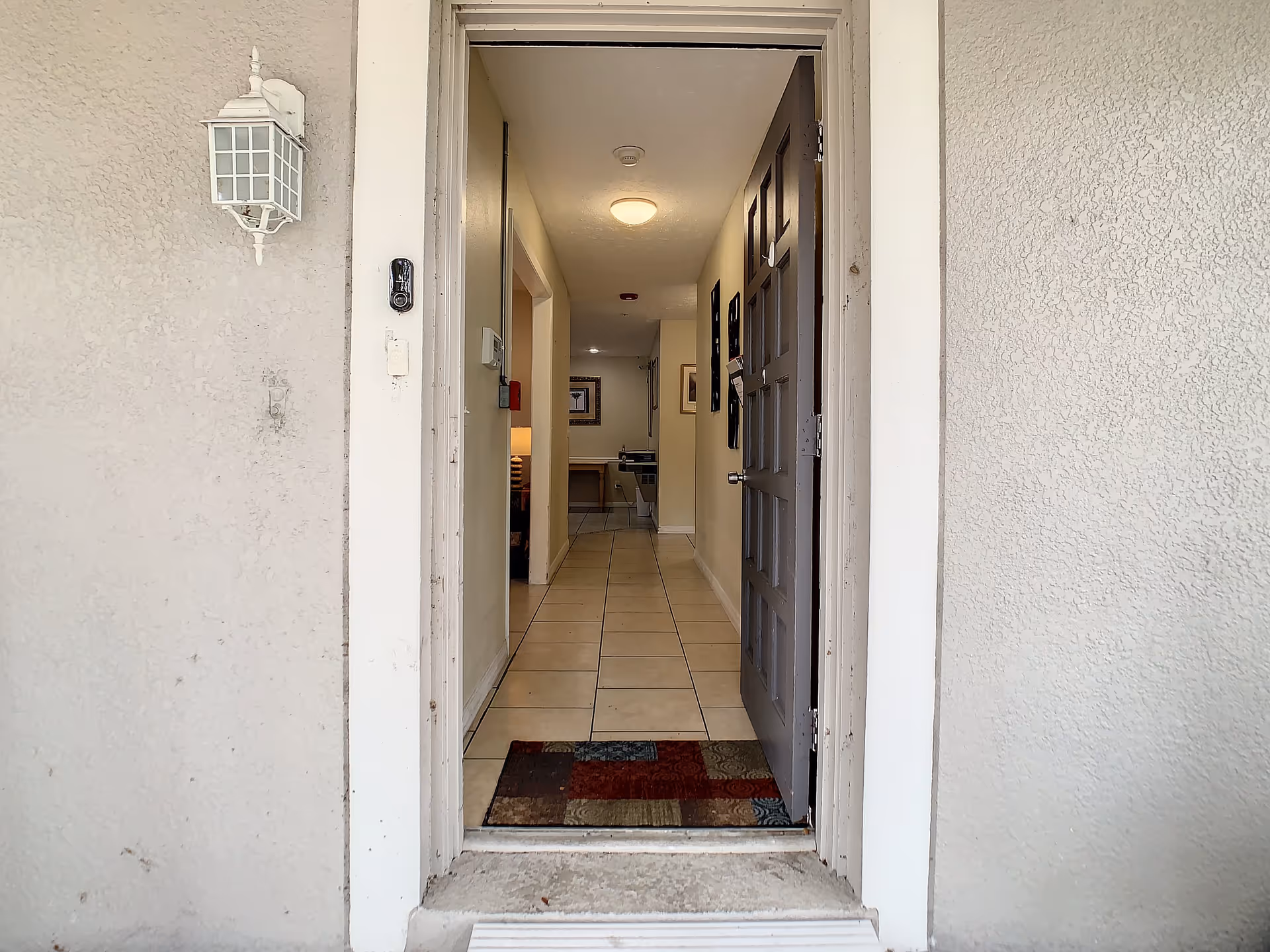 View through an open front door into a hallway with tiled floor and beige walls. A colorful doormat is placed at the entrance. The hallway is lit by ceiling lights and has framed pictures on the walls. A lamp and furniture are visible in a room to the left.