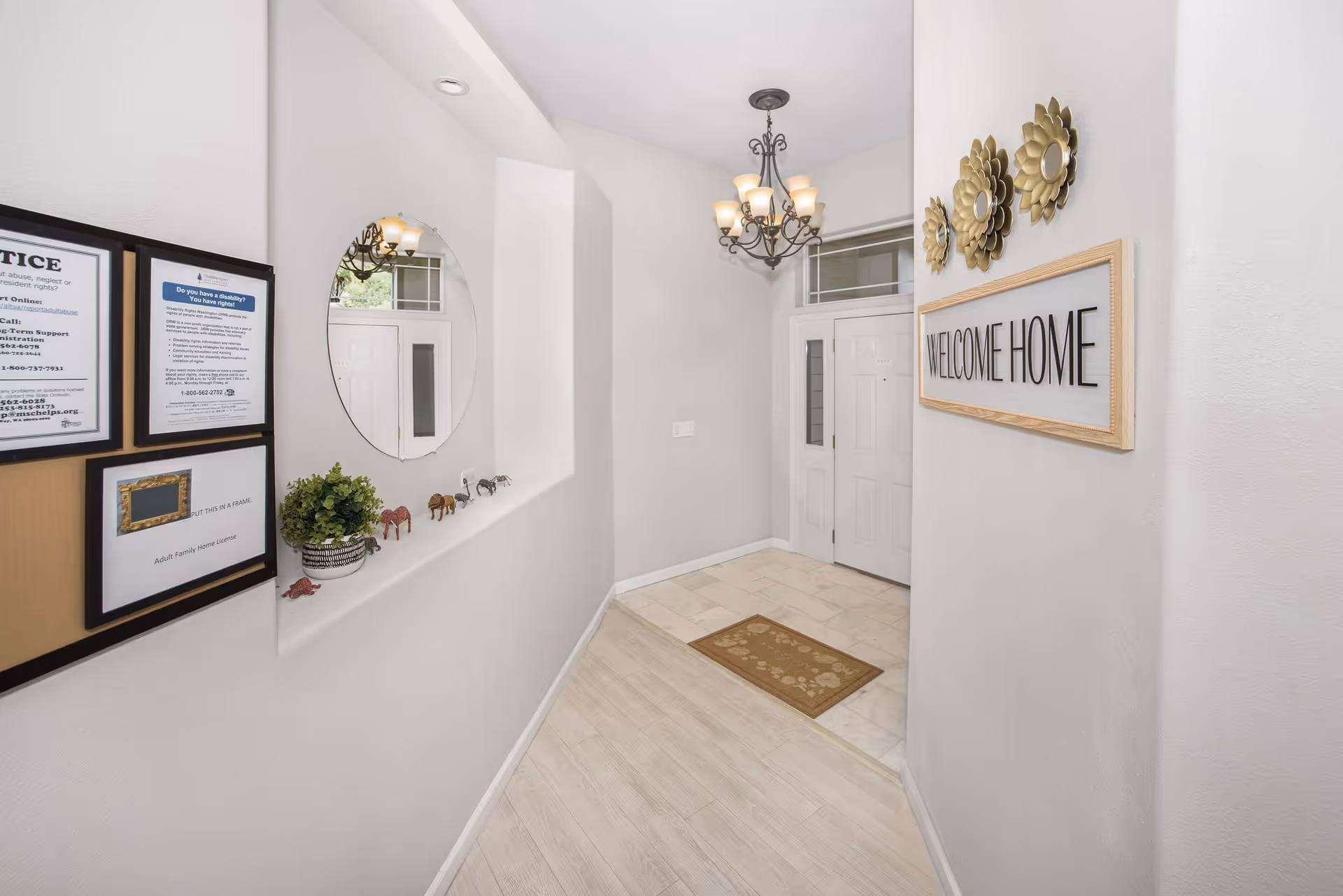 A bright entryway with light-colored wood flooring leading to a white front door with sidelights and a transom window. A decorative chandelier hangs from the ceiling. On the right wall, there is a framed sign that reads 'WELCOME HOME' and three gold flower-shaped wall decorations. On the left wall, there is a round mirror above a ledge with small decorative animal figurines and a potted plant. Below the mirror, there are framed notices and a license displayed.