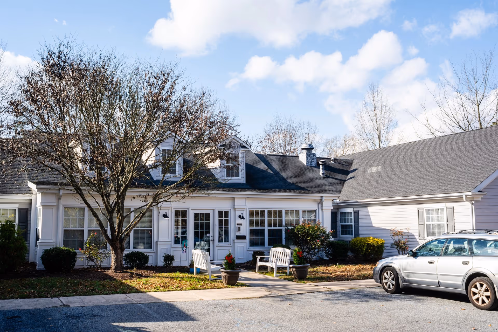 Front exterior of a single-story assisted living building with white siding, a leafless tree, benches, and a parked silver car.