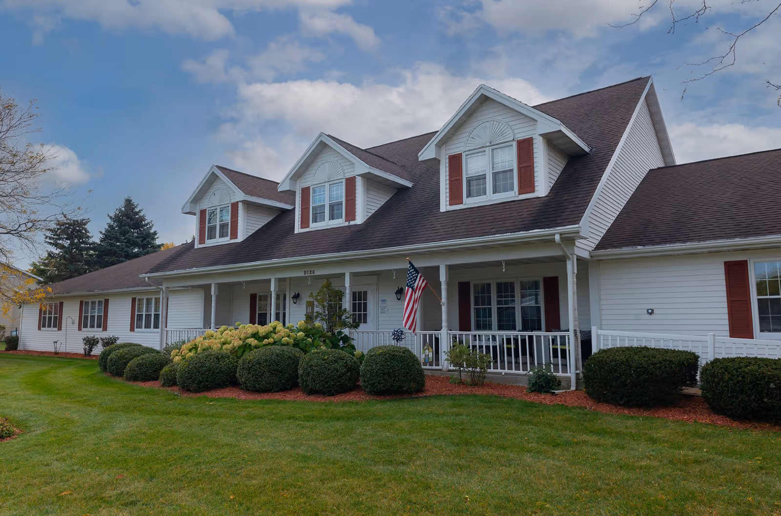 Exterior view of a single-story senior living facility building with white siding, brown roof, and red shutters. The building has a front porch with white railings, an American flag, and well-maintained landscaping with green bushes and grass under a partly cloudy sky.