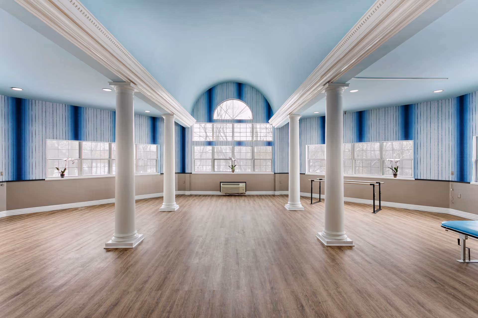 A spacious room with light wood flooring, blue and white striped wallpaper, and large windows letting in natural light. The room features white decorative columns and a high ceiling with crown molding. There are a few pieces of exercise equipment, including parallel bars and a padded bench, suggesting it is used for physical therapy or exercise.