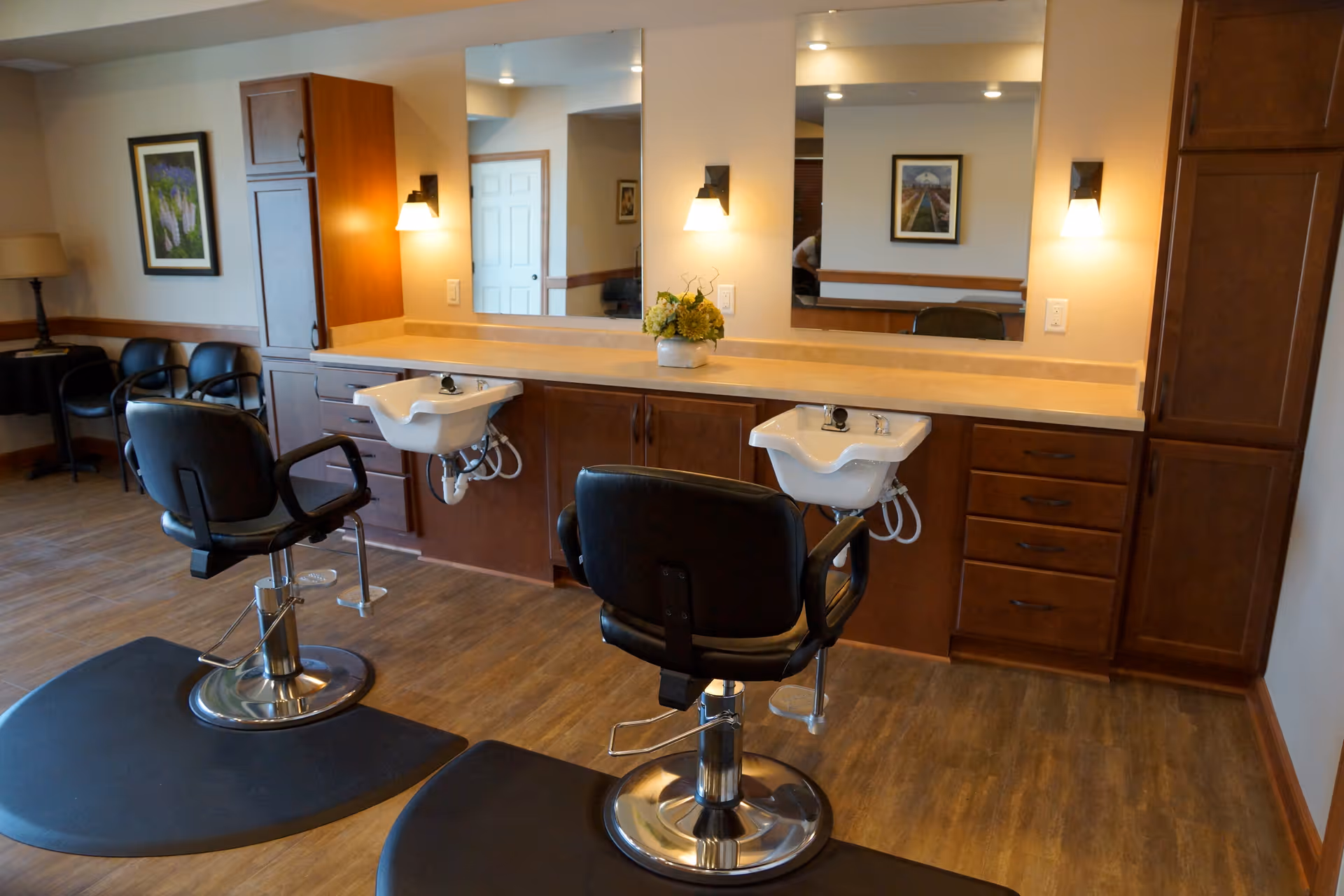 Interior salon area with two styling chairs and shampoo sinks in front of a mirrored counter and wooden cabinets.