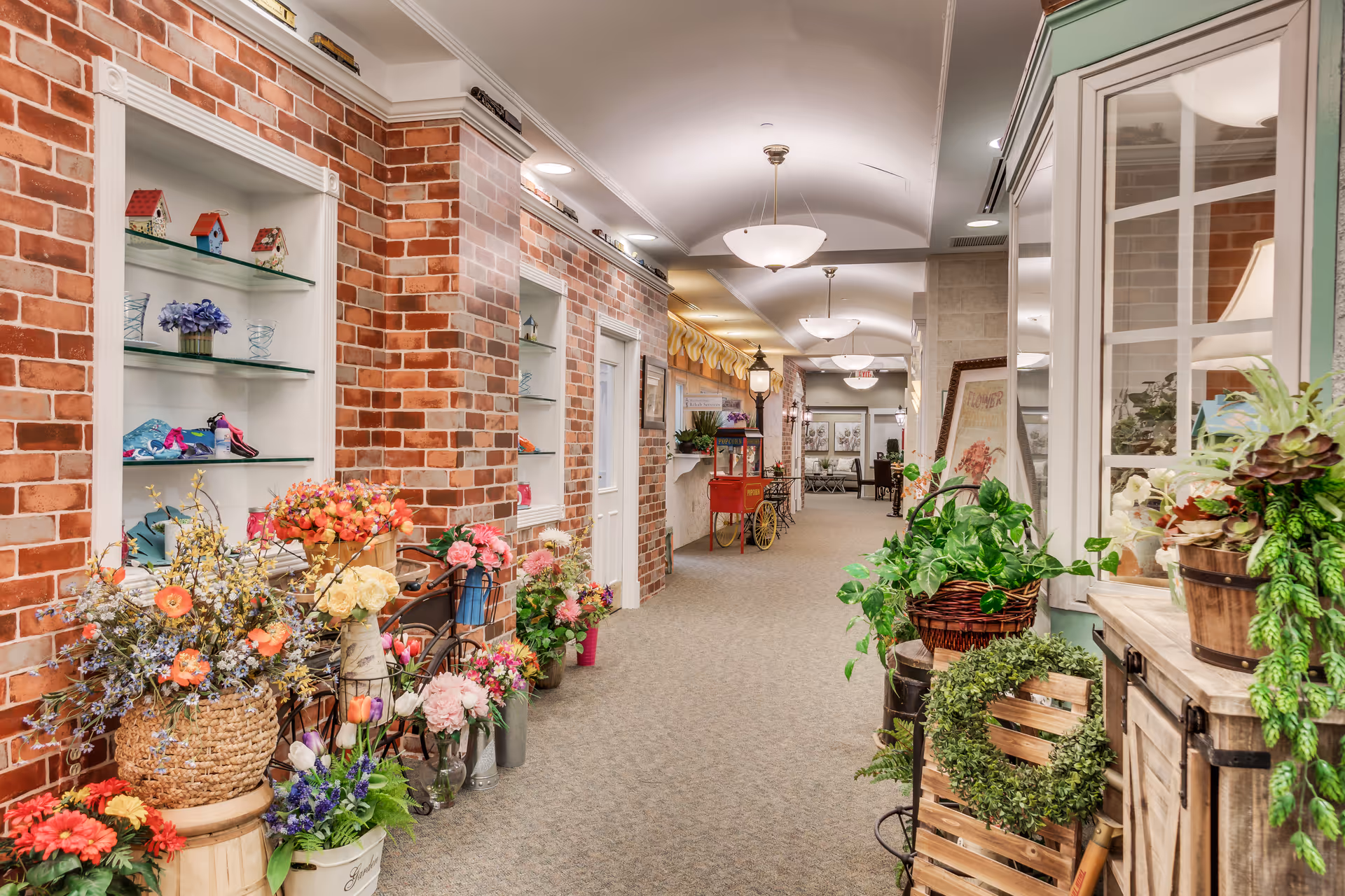 Bright indoor hallway of an assisted living facility decorated with brick storefront facades, potted plants, and floral displays.