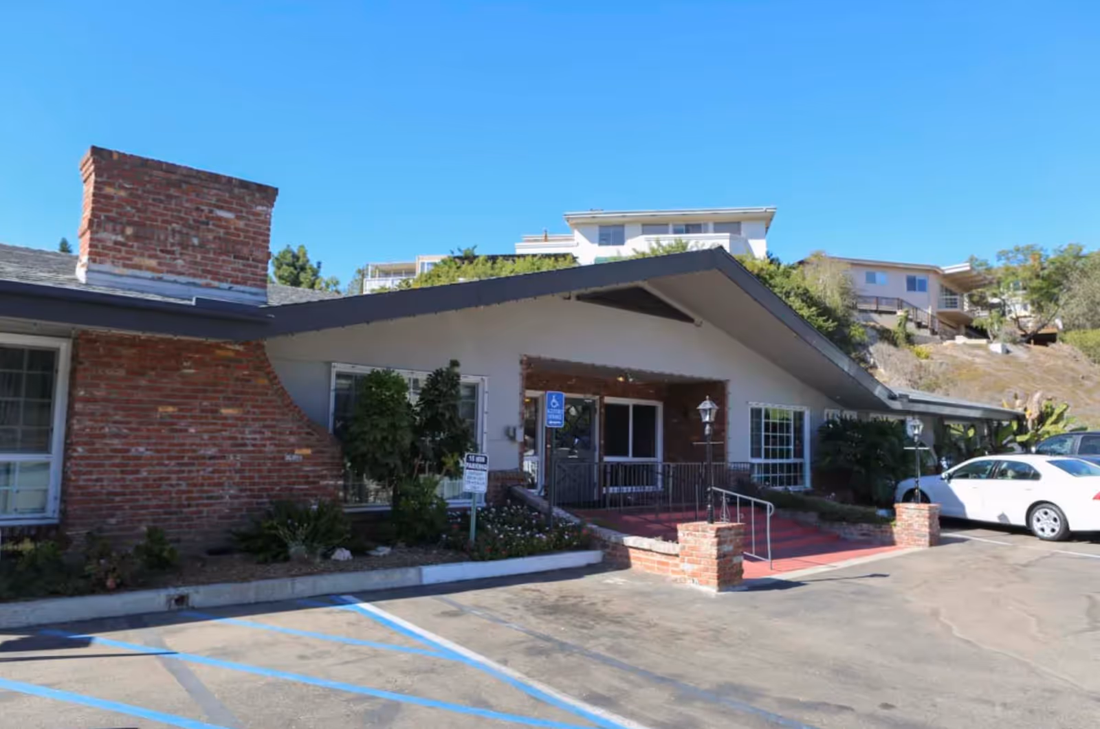 Exterior view of Mission Hills Post Acute Care facility showing a single-story building with a brick chimney, a sloped roof, and a ramp leading to the entrance. There are some plants and bushes near the building, a clear blue sky, and a few parked cars in the parking lot.