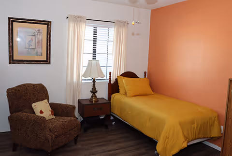 Small bedroom with a single bed covered in yellow bedding against an orange accent wall, a nightstand with a lamp, an upholstered armchair, and a window with curtains.